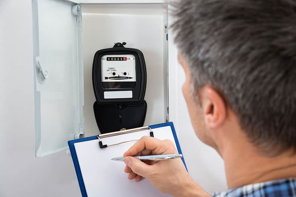 A person records the reading from an electric meter in a wall-mounted box, using a clipboard and pen.