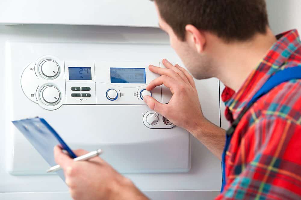 Person in a plaid shirt adjusts controls on a white boiler while holding a clipboard and pen.
