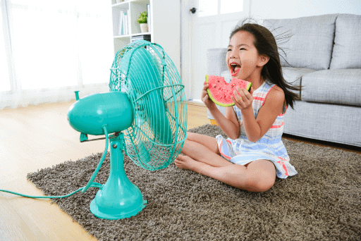 Girl sitting on a carpet, enjoying a slice of watermelon in front of a turquoise fan in a living room.