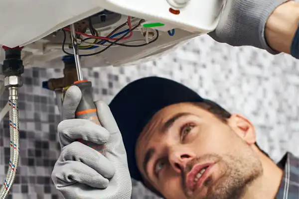 A person wearing gloves and a cap is using a screwdriver to repair electrical components inside a panel, with visible wires and a tiled background.