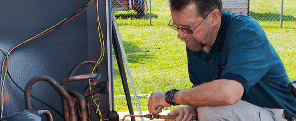 A man is kneeling and examining wiring in an outdoor unit, possibly an air conditioner, with grass and a fence in the background.