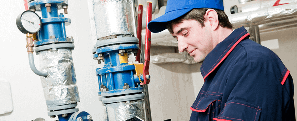 A worker in a blue uniform and cap inspects industrial piping with gauges in a facility.