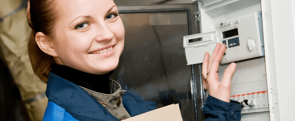 A person in a blue uniform smiles while standing next to an open electrical panel, holding a clipboard and adjusting a device inside.