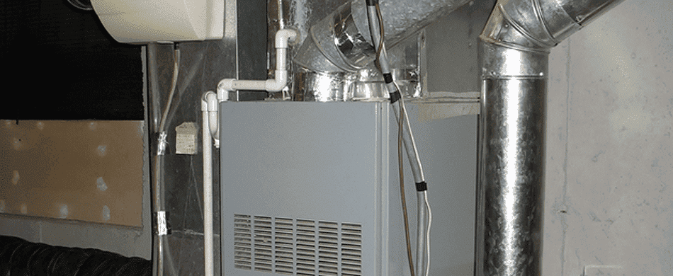 A gray furnace with attached ductwork and pipes in a basement setting. The system is connected to a wall and surrounded by metallic and insulated components.