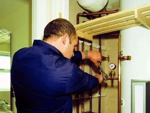 A plumber in a blue uniform is using a wrench to adjust pipes next to a water heater in a utility room.