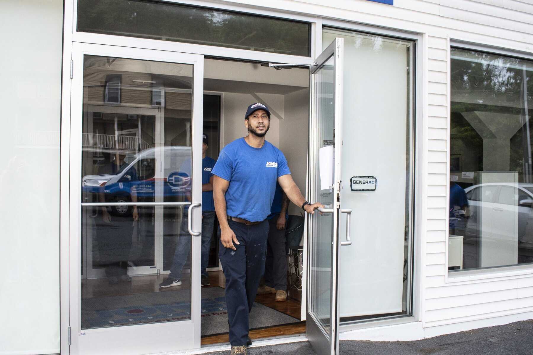 A man wearing a blue shirt and cap opens a glass door of a building. A van is visible through the glass.