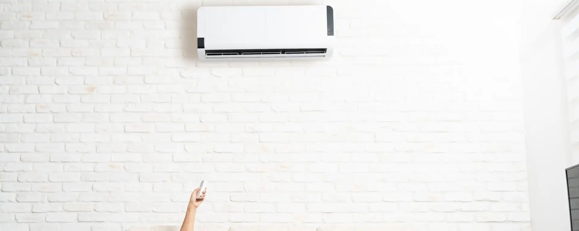 A hand holding a remote points towards a wall-mounted air conditioning unit on a white brick wall.