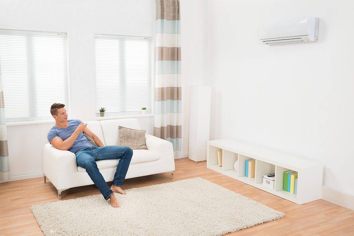 A person sits on a white sofa in a bright room with light wood floors, looking at a wall-mounted air conditioner. A white shelf with books is against the wall.