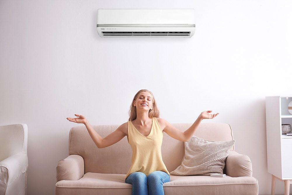 A woman sits on a couch with her arms raised and eyes closed, beneath a wall-mounted air conditioner in a living room setting.