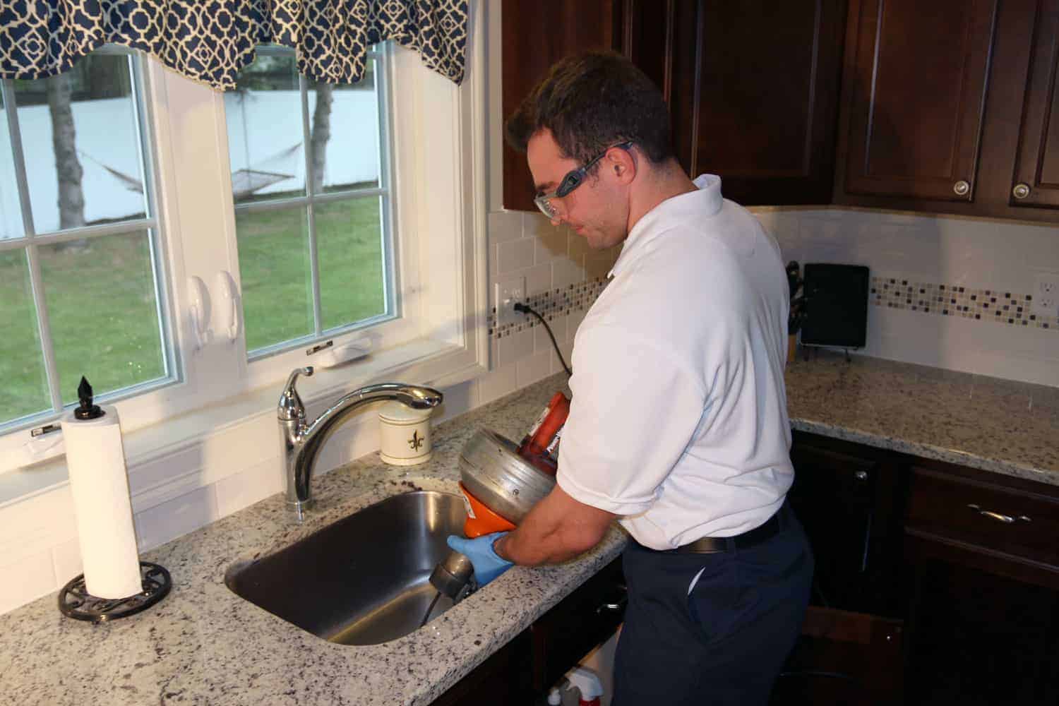 A person wearing gloves is using a plumbing snake to unclog a kitchen sink drain in a home kitchen.