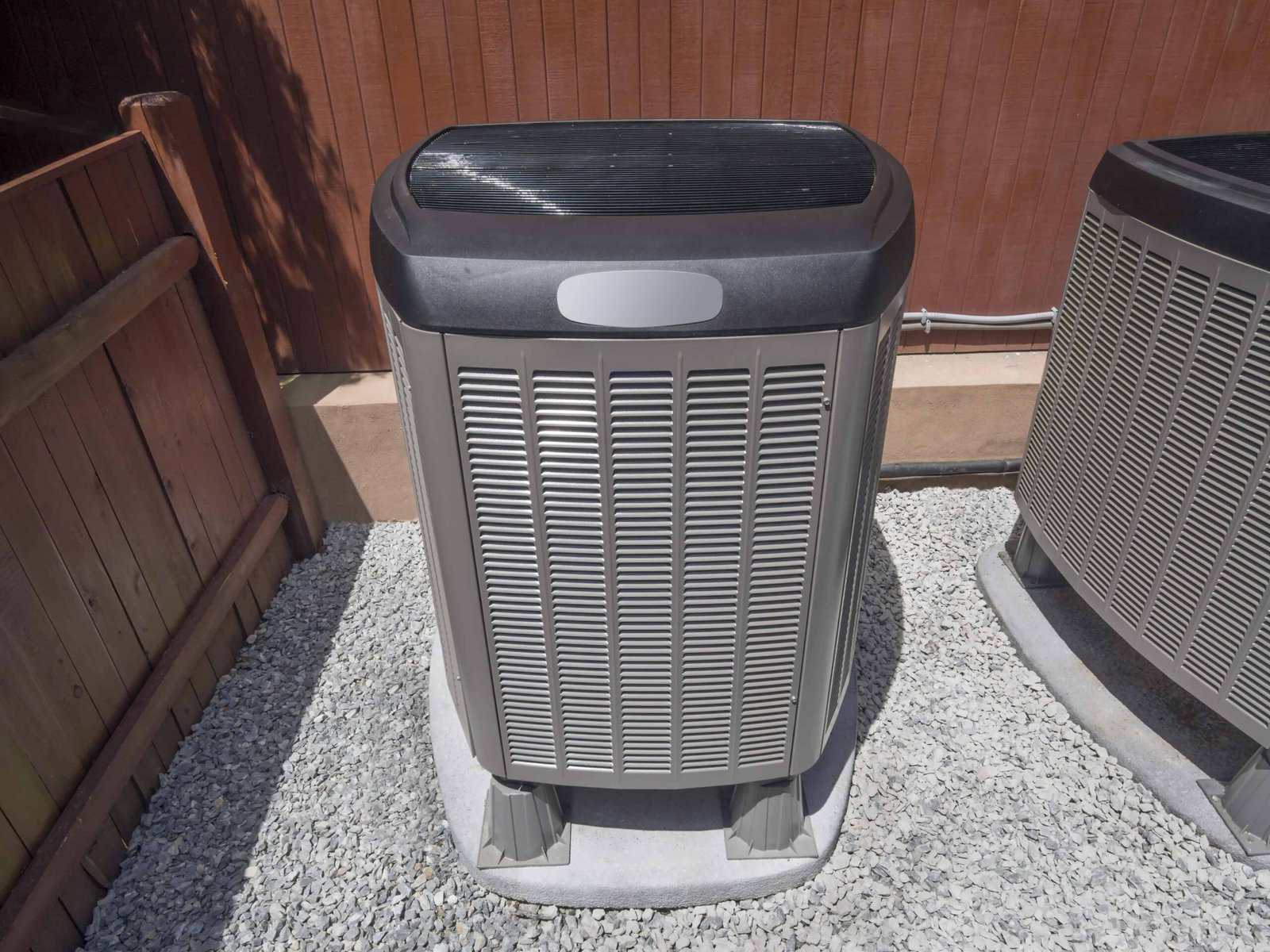 Outdoor air conditioning unit placed on a gravel bed next to a wooden fence and wall.