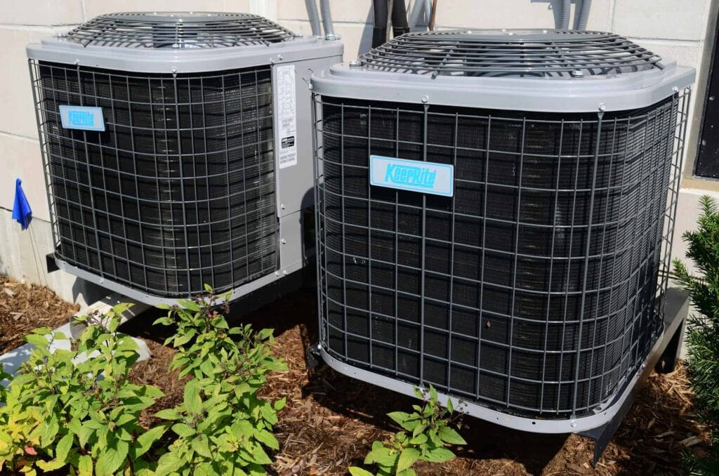 Two outdoor air conditioning units are installed beside a building on a concrete surface, surrounded by green plants and mulch.