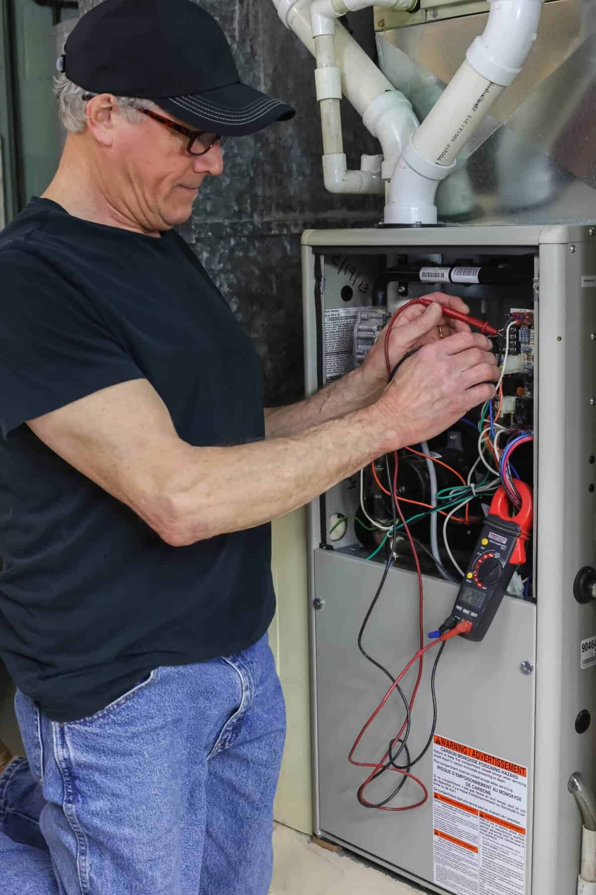Man in black shirt and cap uses a multimeter for wiring diagnostics on an open HVAC unit.