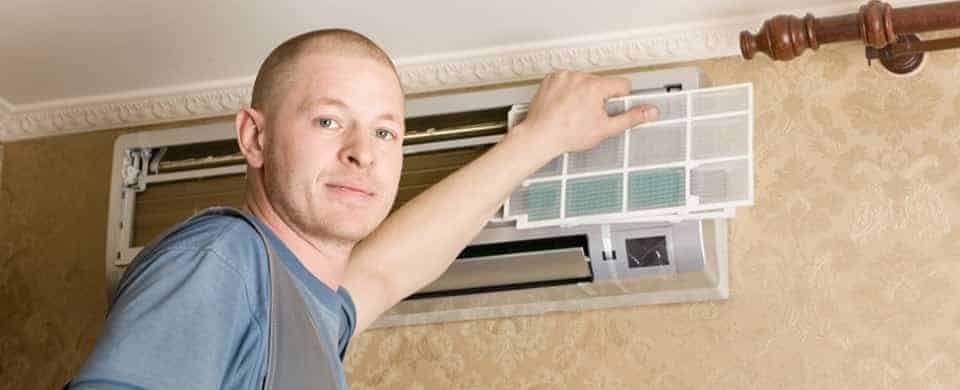 Person in a blue shirt and overalls cleaning or installing an air conditioning unit mounted on a wall.