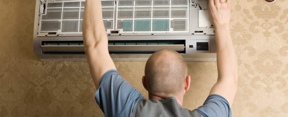 A person with a shaved head and a blue shirt inspects a wall-mounted air conditioning unit inside a room.