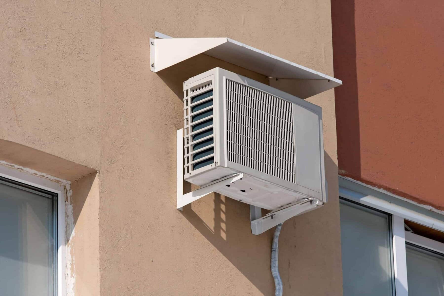 Wall-mounted air conditioning unit outside a building, situated between two windows, with a protective cover on top.