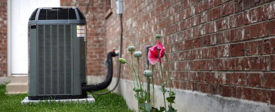 An outdoor air conditioning unit on grass next to a brick wall, with pink flowers growing nearby.