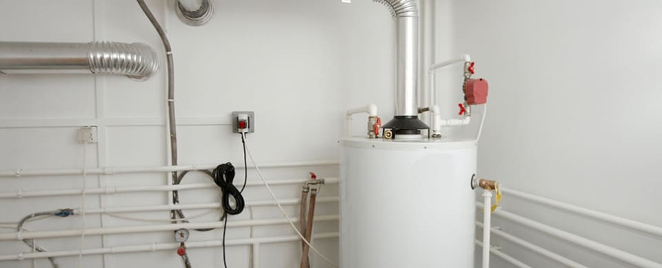 A white water heater is installed in a room with white walls, surrounded by pipes and a ventilation duct. A black hose hangs from a nearby pipe, and a red valve is visible on the heater.