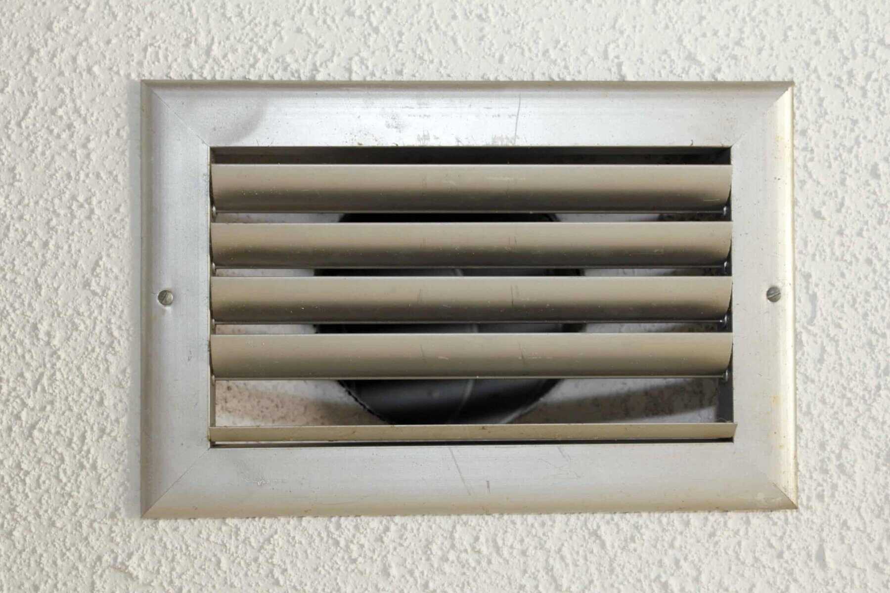 A silver metal air vent with horizontal slats is set into a textured white wall.