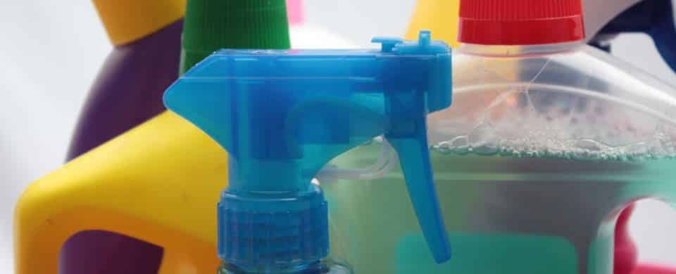 Close-up of various cleaning product bottles, including a blue spray bottle, a purple and yellow bottle, and a clear bottle with red cap, partially filled with liquid.