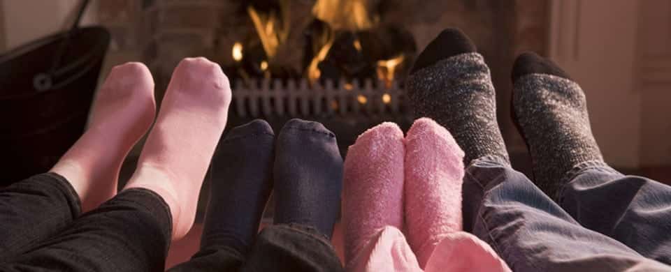 Four pairs of feet in socks are lined up in front of a lit fireplace.