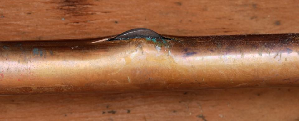 A copper pipe with a visible crack and small bulge, showing signs of wear and corrosion, set against a wooden background.