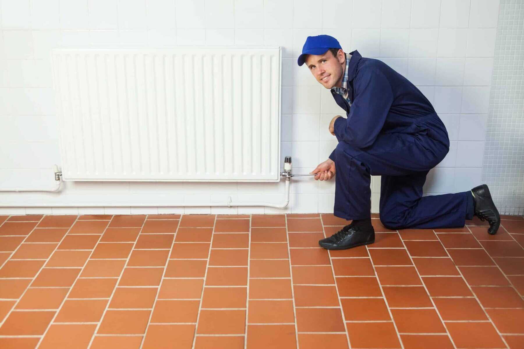 A person in a blue jumpsuit and cap kneels on a tiled floor, adjusting a radiator valve on a white wall.