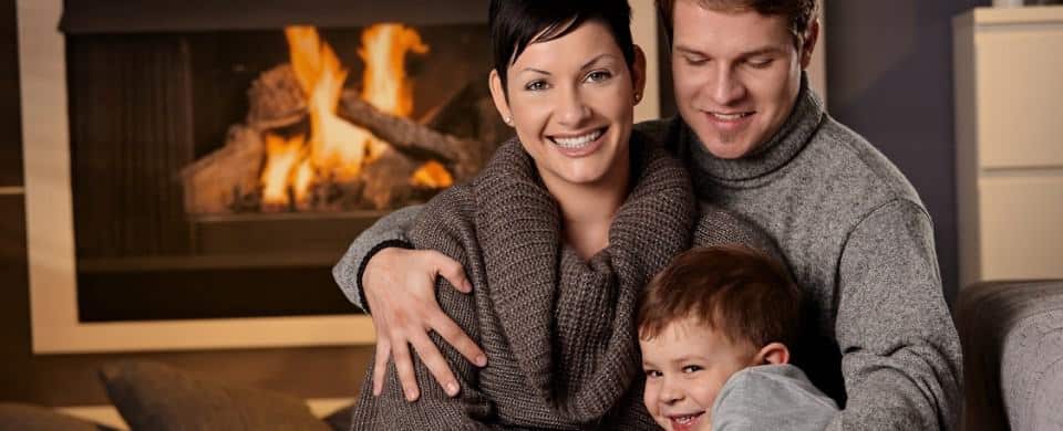 A family of three sits together, smiling, in front of a fireplace.