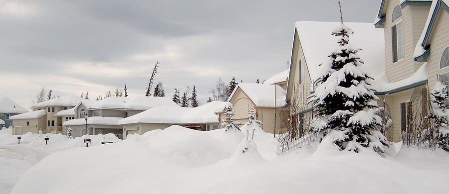 Snow-covered suburban houses and trees under a cloudy sky.