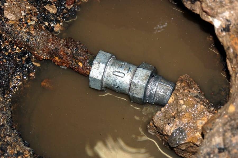 A corroded metal pipe connected with a rusty coupler is partially submerged in muddy water and surrounded by dirt and debris.