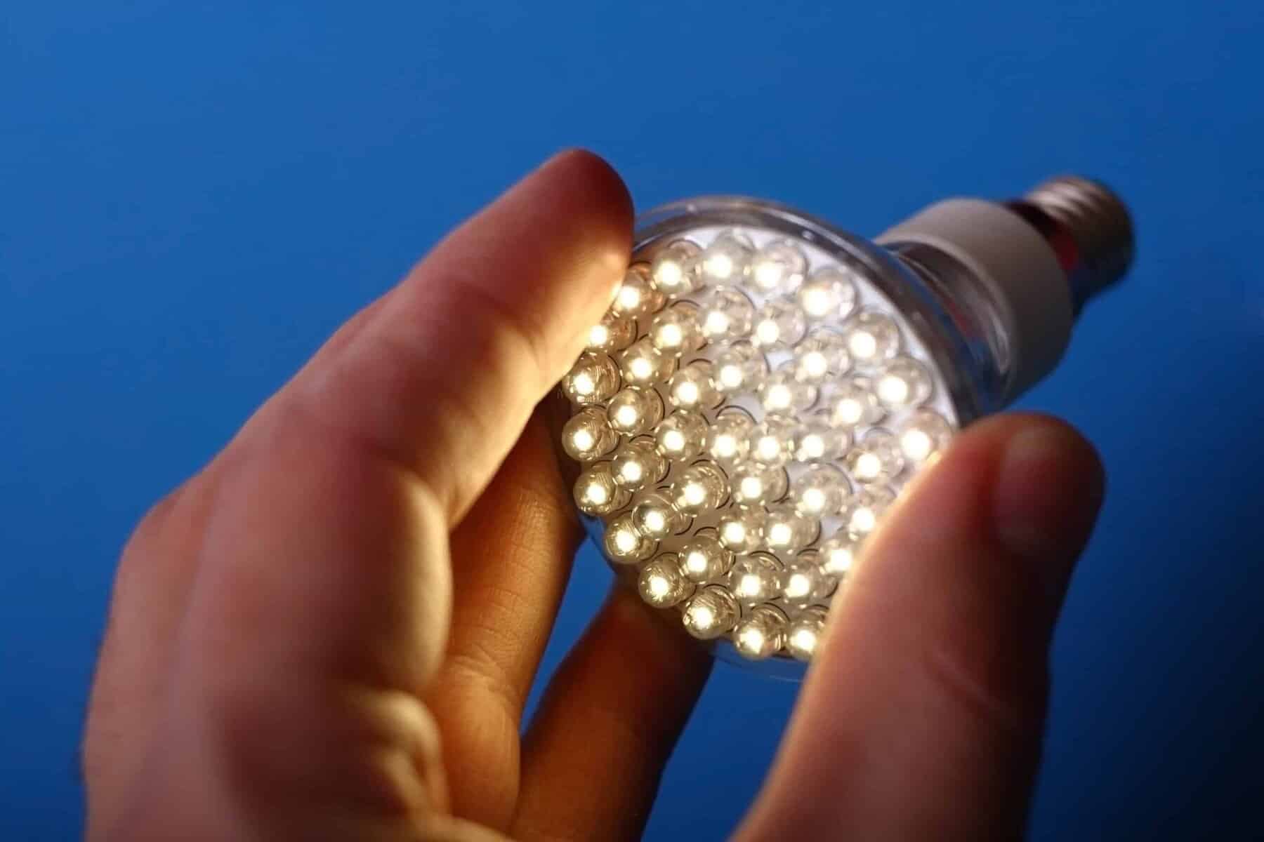 A hand holds an illuminated LED light bulb against a blue background.