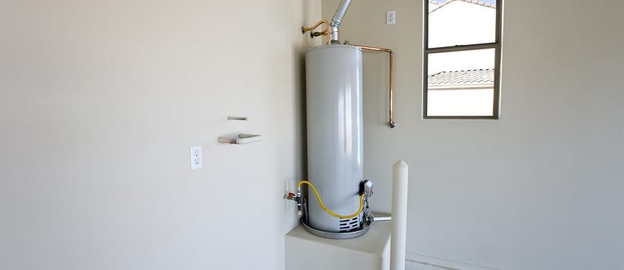 A gray water heater installed in a corner of a room with beige walls and a window.