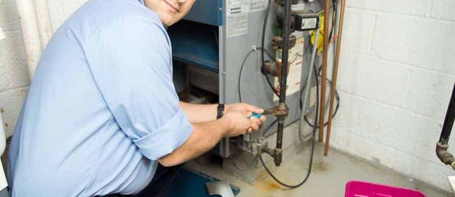 Man in a blue shirt repairing or inspecting a furnace in a basement or utility room.