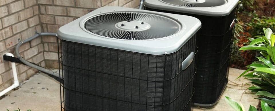 Two outdoor air conditioning units on a concrete slab next to a brick wall, surrounded by green plants.