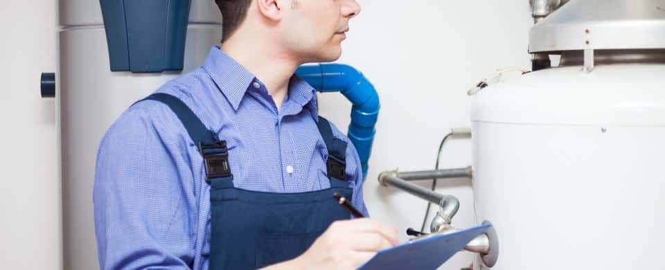 A man in blue overalls examines a water heater, holding a clipboard, in a utility room.