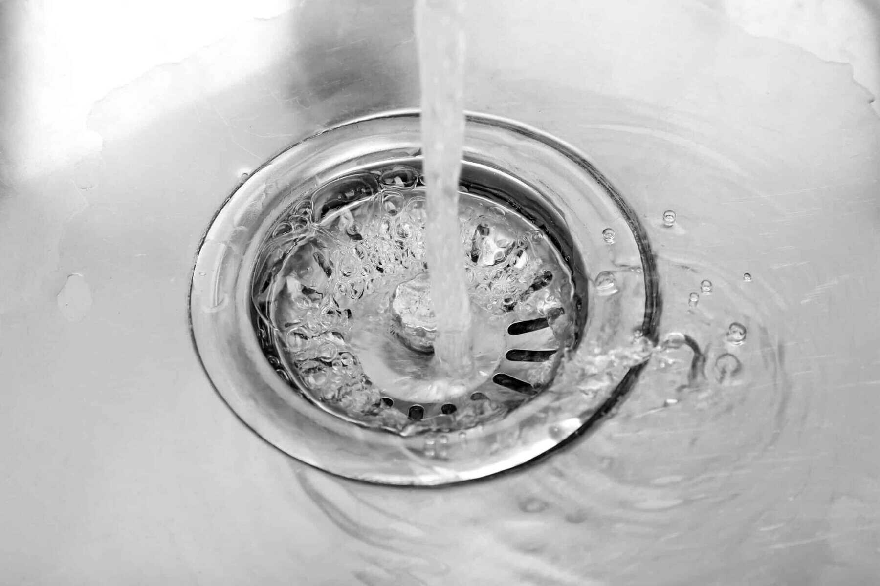 Water flowing from a tap into a stainless steel sink drain, creating ripples and bubbles in the water.