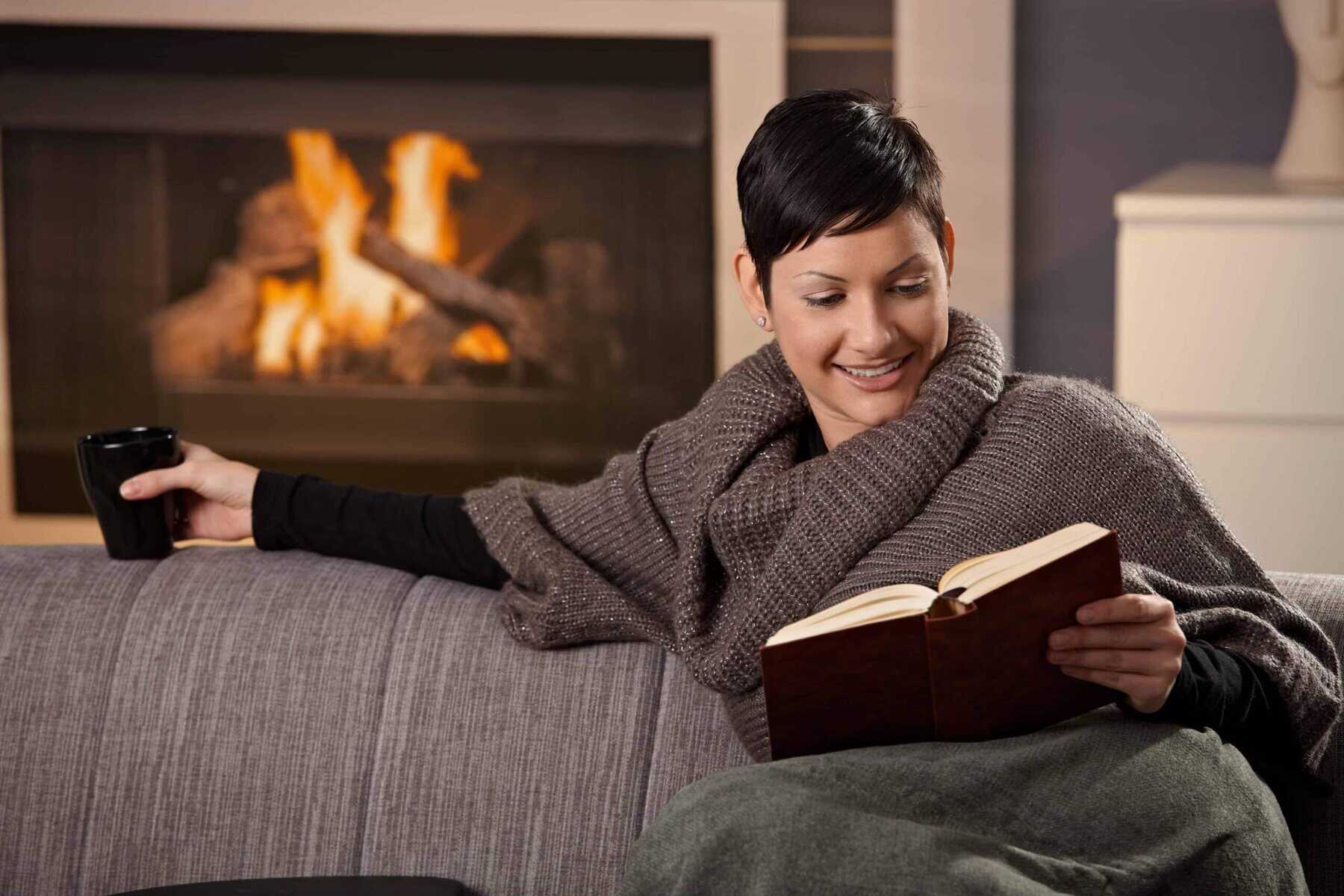 Person sitting on a couch by a fireplace, wrapped in a blanket, holding a mug, and reading a book.