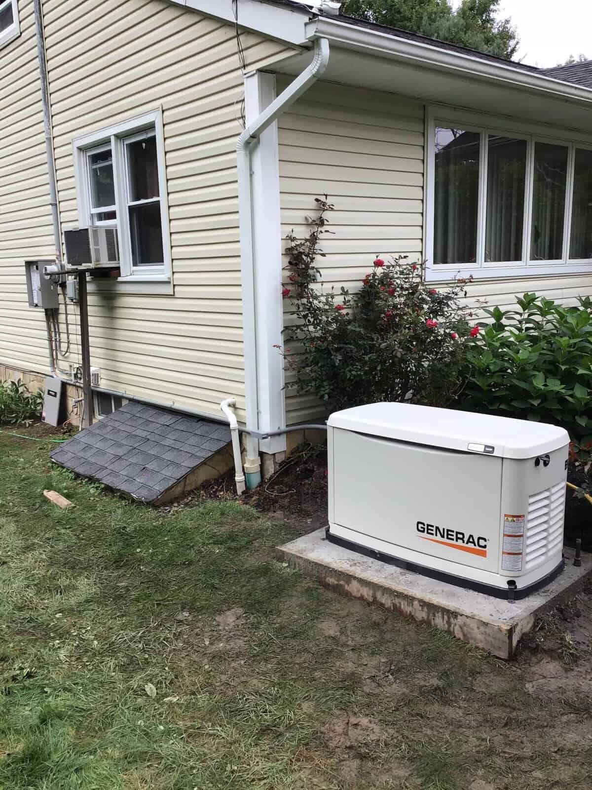 Outdoor generator installed near a house with beige siding, beside a garden with flowers and shrubs. A basement window and an air conditioning unit are visible.