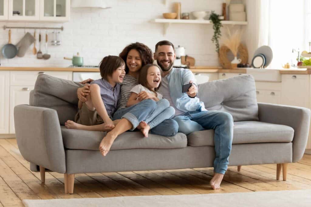 A family of four, sitting on a gray couch, smiling and laughing together in a bright living room with a kitchen in the background.