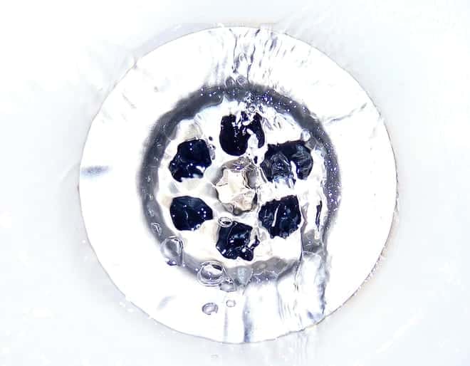 Close-up of a sink drain with silver sides, water droplets, and small black debris caught inside.