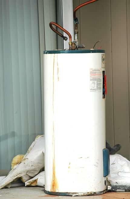 A vertical white water heater with visible rust stains is placed outdoors near a sliding glass door.