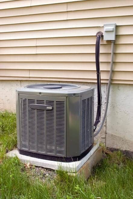 An outdoor air conditioning unit is placed on a concrete slab next to a house with beige siding. Power cables connect the unit to an electrical box on the wall. Grass surrounds the area.
