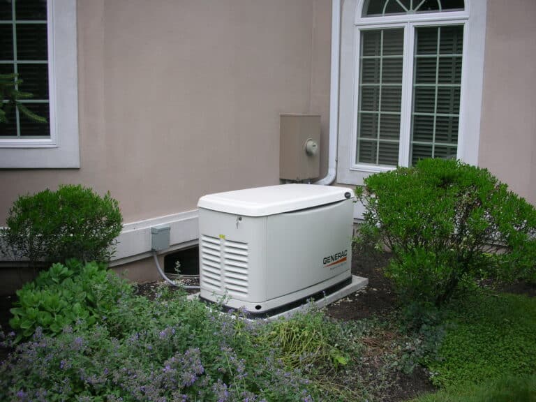 A white backup generator is installed outside a house next to a window and surrounded by bushes and plants, illustrating a neat and professional home generator installation.