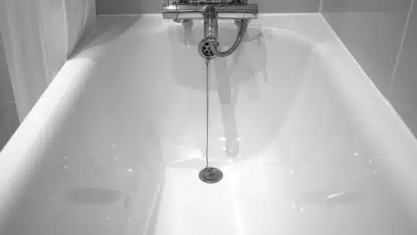 A white empty bathtub with a metal drain plug on a chain, silver faucet, and tiled walls in the background; ideal for showcasing professional Drain Clearing Services.
