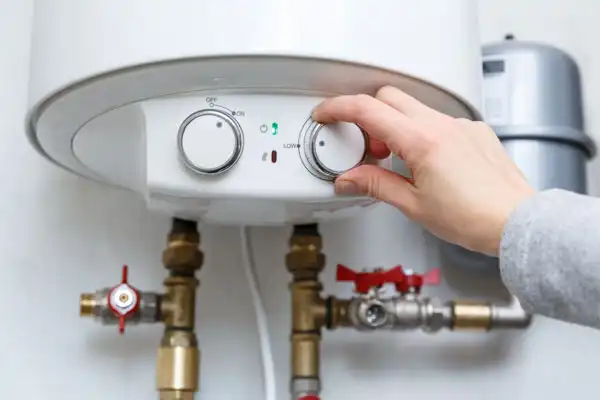 A person adjusts the temperature dial on a white water heater with visible pipes and control panel, checking settings after boiler installation.