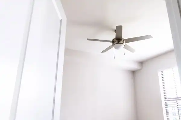 A modern silver ceiling fan with five blades is expertly installed on a white ceiling in a bright, empty room with a window and white walls—perfect for those seeking ceiling fan installation in Goshen NY.