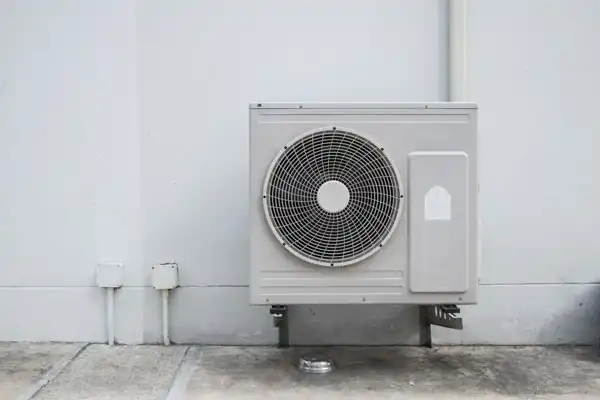 An outdoor heat pump installation is mounted on a wall above a concrete surface, with pipes and electrical connections visible nearby.