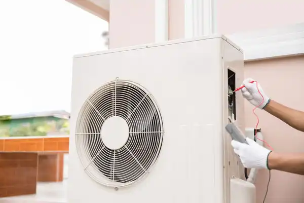 A person wearing white gloves uses a multimeter to check the wiring of an outdoor air conditioning unit during heat pump maintenance in Goshen NY.
