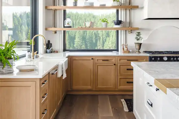 Modern kitchen with light wood cabinets, white countertops, gold faucet, floating shelves with plants and dishes, and a large window overlooking trees—perfect for those who appreciate style and seamless plumbing repair accessibility.