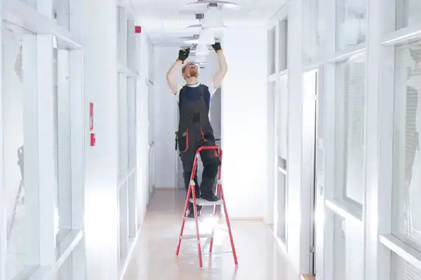 A worker stands on a red step ladder performing lighting repair, installing or fixing a ceiling light fixture in a bright hallway with glass-paneled walls.